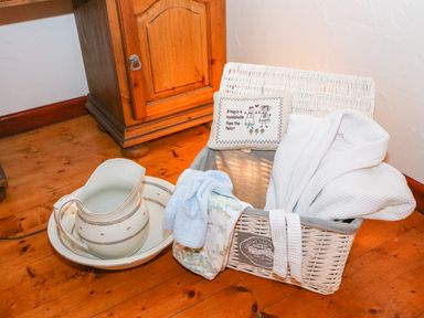 A wicker basket with towels and a bathrobe next to a ceramic water pitcher and basin on a wooden floor at Woodleigh Cottage in Ballycastle