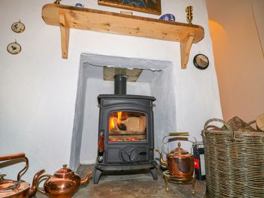 A wood burning stove with fire inside surrounded by copper kettles and a basket of firewood at Woodleigh Cottage in Ballycastle