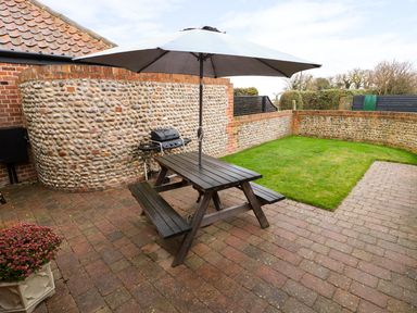 An outdoor patio with a wooden picnic table and umbrella a barbecue grill and a small grassy area at Hitchens Cottage in Happisburgh