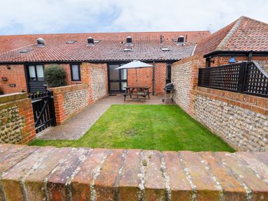 A small enclosed backyard with grass a paved seating area a wooden picnic table with an umbrella and a barbecue at Hitchens Cottage in Happisburgh
