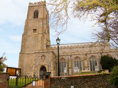 An old stone church with a tall bell tower surrounded by trees and a fence at Hitchens Cottage in Happisburgh