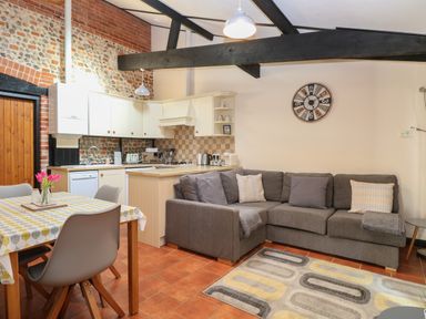 An open kitchen and living area with a dining table with chairs a gray sectional sofa and a wall clock at Hitchens Cottage in Happisburgh