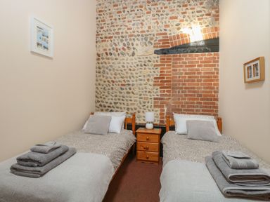 A bedroom with two single beds a wooden nightstand with a lamp and stone and brick wall at Hitchens Cottage in Happisburgh