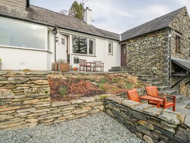 An outdoor stone patio area with wooden chairs and a stone house with white and stone walls at Kirkstone Cottage in Outgate near Hawkshead