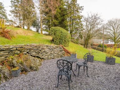 A patio area with black metal table and chairs on gravel surrounded by grass and stone wall at Kirkstone Cottage in Outgate near Hawkshead