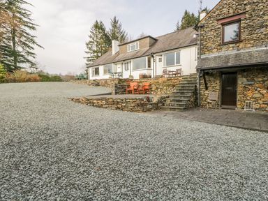 A gravel driveway leading to a stone and white house with outdoor benches and stone stairs at Kirkstone Cottage in Outgate near Hawkshead
