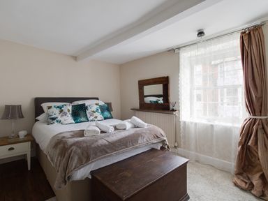 A bedroom with a bed, decorative pillows, bedside table with lamp and cup, wooden chest, mirror on radiator, and window with curtains at Fenders Brewers Quay Harbour