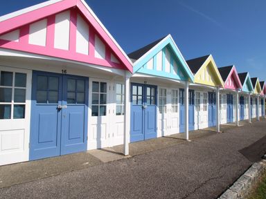 A row of beach huts with blue doors and pink blue and yellow roofs along a paved walkway