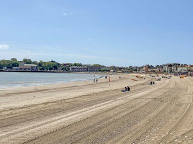 A sandy beach with people sitting and walking near the sea and buildings in the background at Fenders in Brewers Quay Harbour