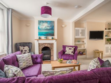 A living room with purple sofas and armchairs a fireplace with painting above and a wall-mounted television at Wey Cottage in Weymouth