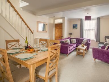 A dining area with a wooden table set for four and a living room with purple sofas at Wey Cottage in Weymouth