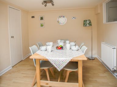 A dining room with a wooden table covered by a polka dot tablecloth six gray chairs white bowls and cups a floor lamp and a wall clock at Wey Cottage in Weymouth
