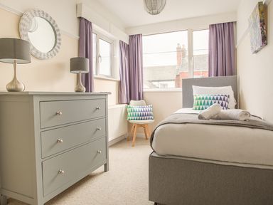 A small bedroom with a single bed a grey dresser two lamps a round mirror a chair and purple curtains at Wey Cottage in Weymouth