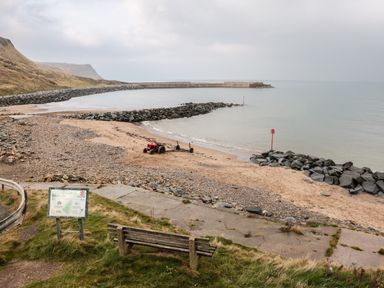 A bench and informational sign near a rocky beach with a small red tractor and breakwater at Lady Bird Retreat in Skinningrove