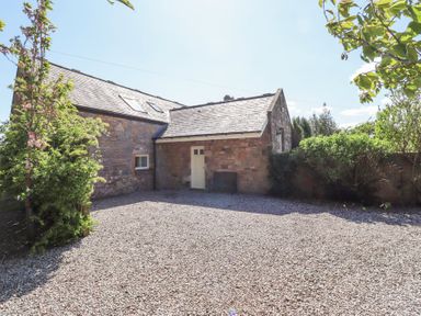 A gravel driveway in front of a stone building with a sloped roof and white door surrounded by bushes at The Coach House in Lowick near Holy Island