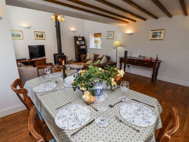 A dining table set with plates glasses and cutlery next to a living area with a wood burning stove and wooden beams on the ceiling at The Coach House in Lowick near Holy Island