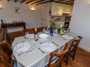 A dining table set for six with plates wine glasses cutlery and flowers in a room with wooden chairs and a kitchen in the background at The Coach House in Lowick near Holy Island