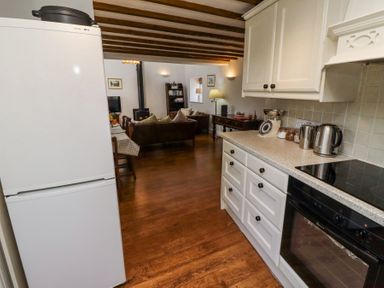 A kitchen area with white cabinets and appliances leading to a living room with a wood stove and brown sofas at The Coach House in Lowick near Holy Island