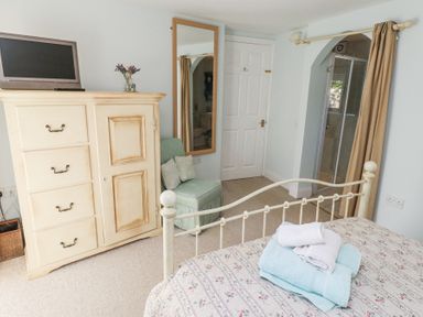 A bedroom with an iron bed frame and floral bedspread with towels on top a cream wardrobe with a TV on it a green chair and a tall mirror in the corner at The Coach House in Lowick near Holy Island