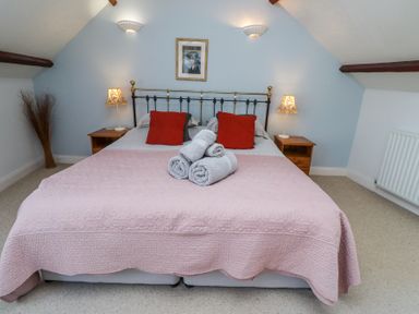 A bedroom with a double bed with pink bedspread and red cushions two bedside tables with lamps and rolled towels on the bed at The Coach House in Lowick near Holy Island