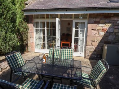 An outdoor patio with a glass table and six checked cushioned chairs outside a stone building with white framed French doors at The Coach House in Lowick near Holy Island