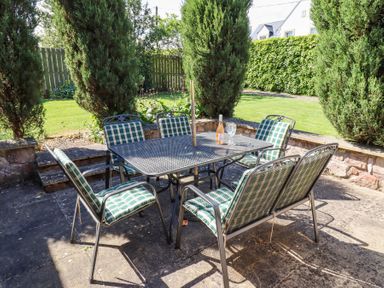 An outdoor patio area with a metal table and six chairs with green checkered cushions surrounded by trees and a grass lawn at The Coach House in Lowick near Holy Island