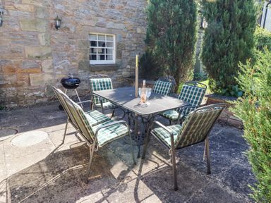 A patio with a metal table and six cushioned chairs with a bottle and two glasses on the table at The Coach House in Lowick near Holy Island