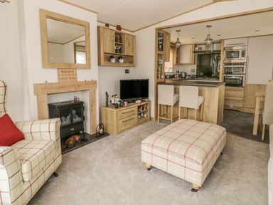 A living room with a plaid armchair and ottoman a TV on a wooden stand a fireplace and an open kitchen with bar stools at 27 Lake View in Cawston