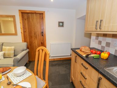 A kitchen with wooden cabinets, countertop with bread and vegetables, a dining table with dishes, and a sofa with cushions at Yr Hen Stabal in Trawsfynydd