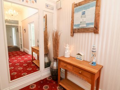 A hallway with a wooden console table holding decorative lighthouses and a calendar next to a tall vase with dried branches and a large wall mirror at Babbacombe Hall in Babbacombe