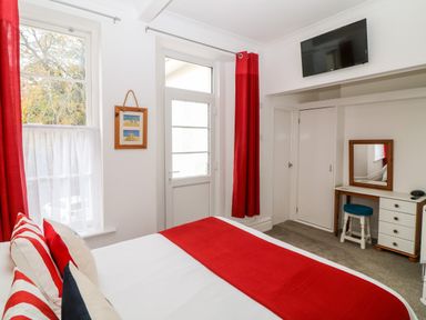 A bedroom with a bed with red and white bedding red curtains a door two windows a wall-mounted television and a dressing table with a mirror at Babbacombe Hall in Babbacombe