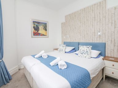 A bedroom with two beds pushed together with blue cushions white pillows and rolled white towels on a blue runner with wallpaper behind and a framed painting on the wall at Babbacombe Hall in Babbacombe