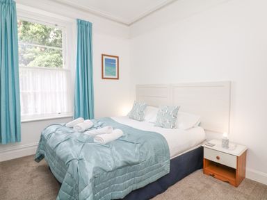 A bedroom with a double bed blue bedding and pillows bedside table with lamp and window with blue curtains at Babbacombe Hall in Babbacombe