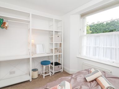 A small bedroom with open white shelving, a stool with a blue seat, a lamp, a mirror, and a window with sheer curtains at Babbacombe Hall in Babbacombe