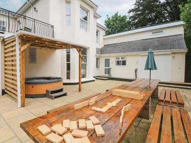 An outdoor patio area with a wooden table and benches a hot tub under a wooden pergola and a house in the background at Babbacombe Hall in Babbacombe