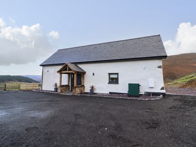 A single-story white building with a slate roof and a wooden porch in a rural landscape at Tyn Llwyn Barn in Glyndyfrdwy near Llangollen
