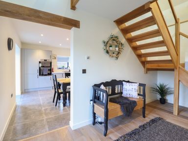 A wooden bench with cushions and a throw under a wooden staircase next to a dining table in a kitchen at Tyn Llwyn Barn in Glyndyfrdwy near Llangollen