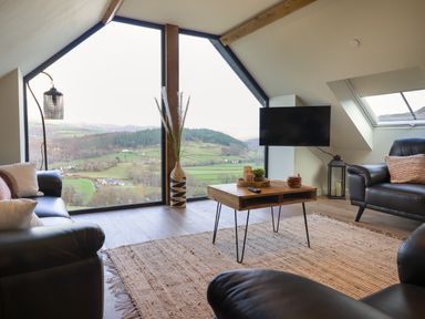 A living room with leather armchairs and a wooden coffee table in front of large windows showing a countryside view at Tyn Llwyn Barn in Glyndyfrdwy near Llangollen