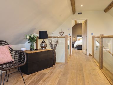 A hallway with wooden flooring glass and wood railings a black chest with lamp and flowers and a wicker chair with a pink cushion at Tyn Llwyn Barn in Glyndyfrdwy near Llangollen