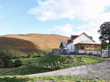 A countryside house on a grassy hill with a fenced yard and a dirt road in front at Tyn Llwyn Barn in Glyndyfrdwy near Llangollen