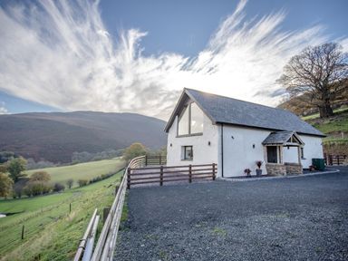 A white barn with a slate roof and a fenced gravel driveway on a hillside with mountains in the background at Tyn Llwyn Barn in Glyndyfrdwy near Llangollen