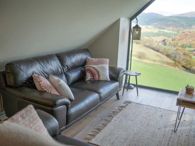 A sitting area with a black leather sofa and pillows beside a large window showing a green landscape at Tyn Llwyn Barn in Glyndyfrdwy near Llangollen