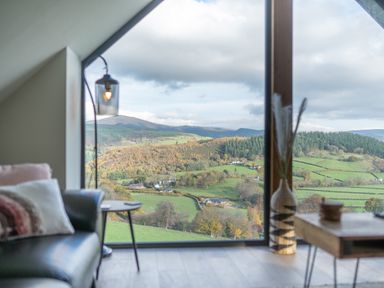 A room with a leather sofa a side table a hanging lamp a wooden coffee table and a large window overlooking hills and fields at Tyn Llwyn Barn in Glyndyfrdwy near Llangollen