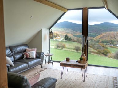 A living room with leather sofas a wooden coffee table and large windows showing a mountain view at Tyn Llwyn Barn in Glyndyfrdwy near Llangollen