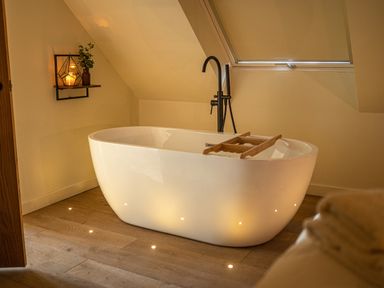 A bathroom with a white freestanding bathtub under a slanted ceiling and a shelf with a candle and plant at Tyn Llwyn Barn in Glyndyfrdwy near Llangollen