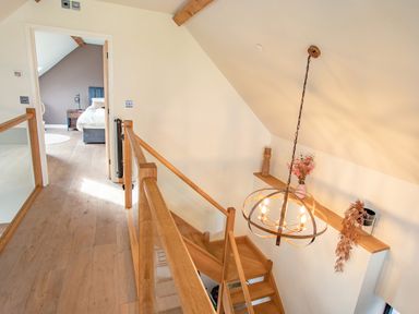 A wooden staircase with a railing and hanging light fixture leading to a hallway with wooden flooring and a bedroom visible at the end at Tyn Llwyn Barn in Glyndyfrdwy near Llangollen