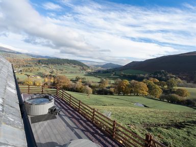 A balcony with a wooden fence and hot tub overlooking a green valley and hills at Tyn Llwyn Barn in Glyndyfrdwy near Llangollen