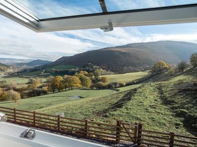 A view of a rural hillside with grass trees and a wooden fence from an open window at Tyn Llwyn Barn in Glyndyfrdwy near Llangollen