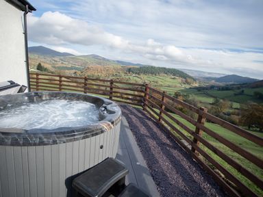 A hot tub on a deck with a wooden railing overlooking a green valley and hills at Tyn Llwyn Barn in Glyndyfrdwy near Llangollen