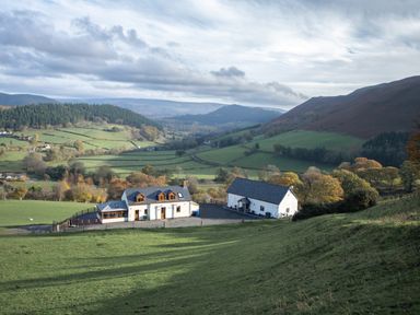 A rural landscape with two white buildings on a green hillside surrounded by fields and hills at Tyn Llwyn Barn in Glyndyfrdwy near Llangollen